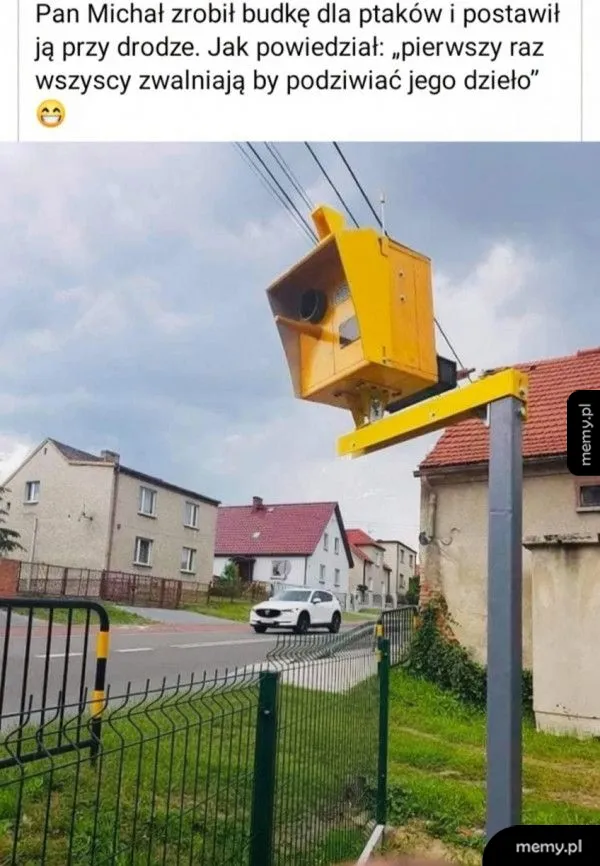 Żółta budka na słupie przy drodze, obok ogrodzenie i domy.