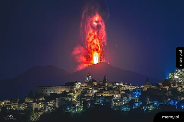 Erupcja wulkanu nocą nad oświetlonym miasteczkiem z widoczną postacią w dymie