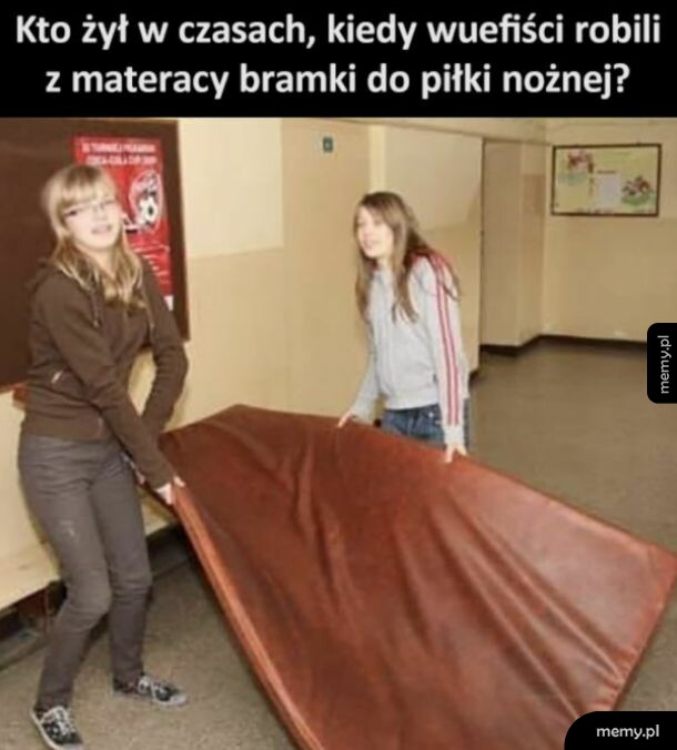 Dwie dziewczynki w szkole niosą duży brązowy materac gimnastyczny.